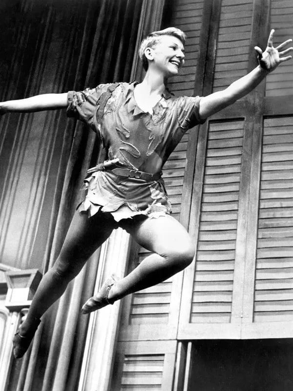 black & white photo of petite, short-haired woman suspended by wires in front of window. She looks like a young boy leaping.